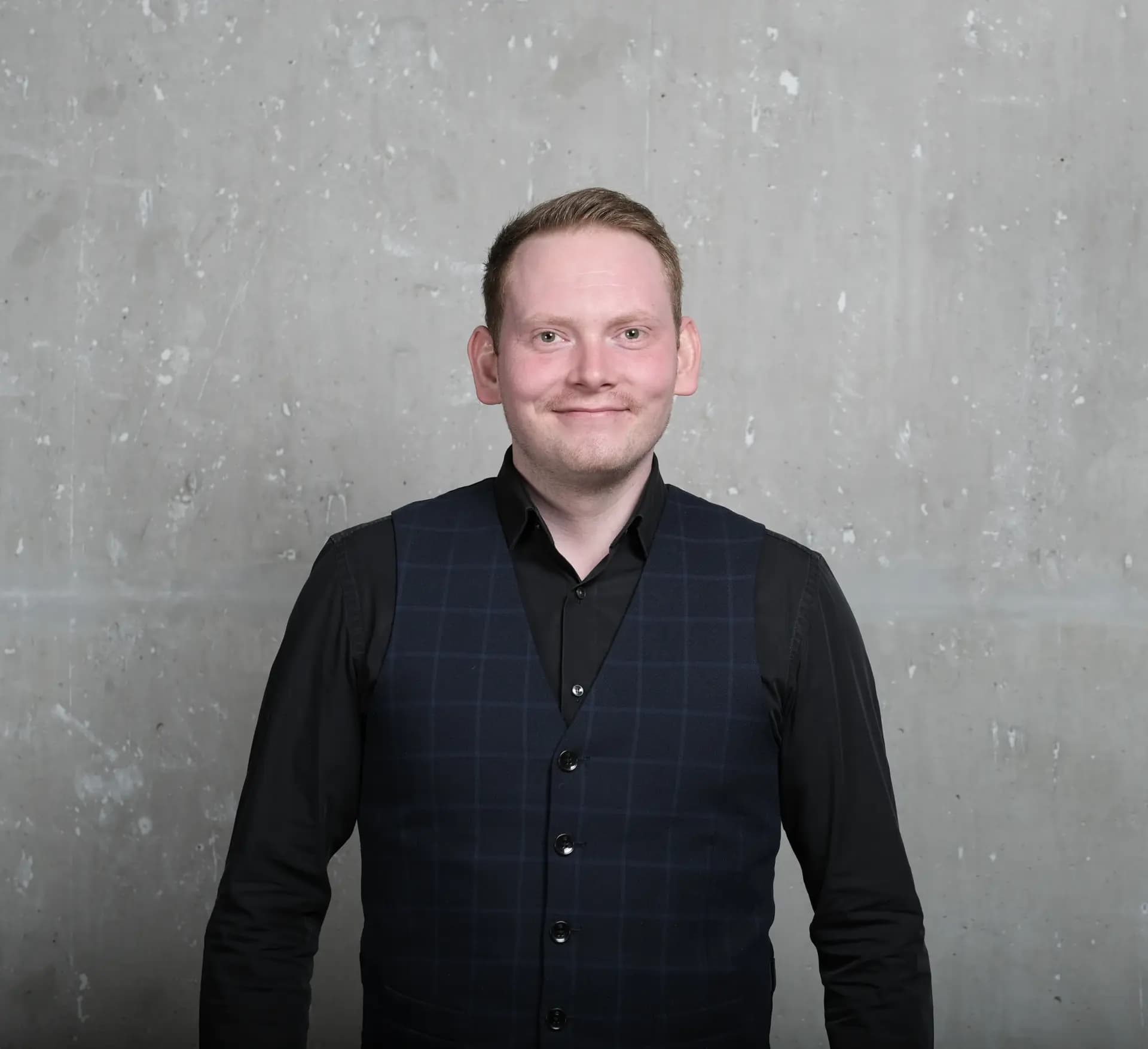 Maximilian Eckel, Gründer und Geschäftsführer von BespokeCode, vor einer grauen Betonwand. Er trägt ein schwarzes Hemd mit einer blau karierten Weste.